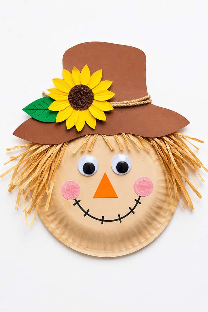 A paper plate face with googly eyes, an orange triangle nose, pink circle cheeks, a stitched black smile, raffia straw hair on the sides, topped with a brown paper hat decorated with a yellow paper sunflower and a green leaf