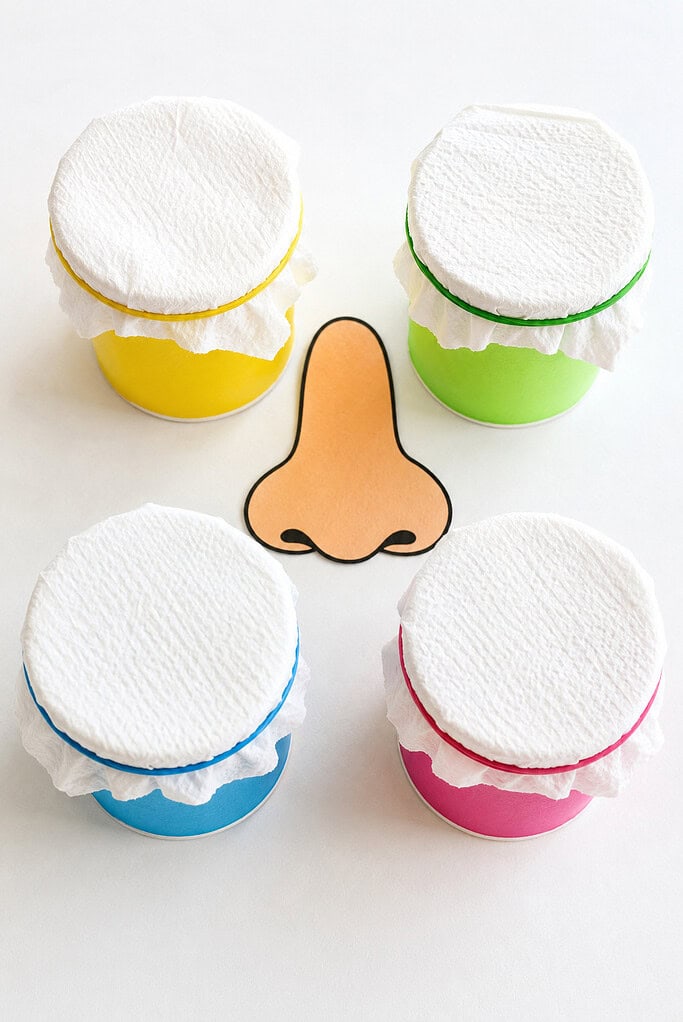 Four colorful paper cups covered with white paper towels and rubber bands, arranged around a paper nose cutout on a bright white table.
