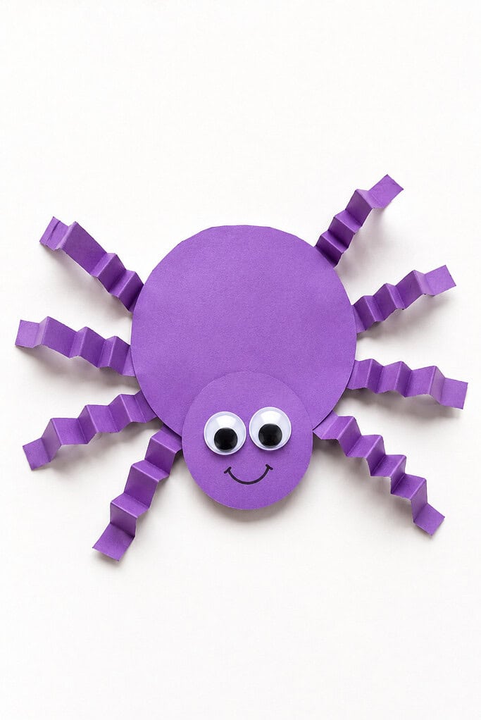 Purple paper spider with a round body and head, googly eyes, a smile, and accordion-folded purple paper legs on a white background