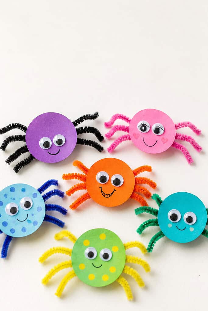 Six colorful pom-pom spiders with googly eyes and pipe cleaner legs in purple, pink, orange, blue, green, and red arranged on a white background