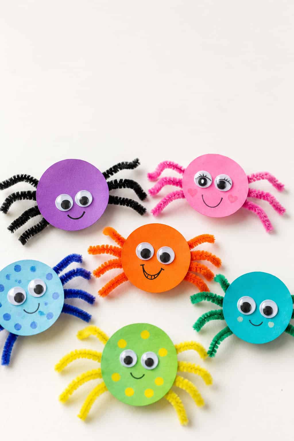 Six colorful pom-pom spiders with googly eyes and pipe cleaner legs in purple, pink, orange, blue, green, and red arranged on a white background