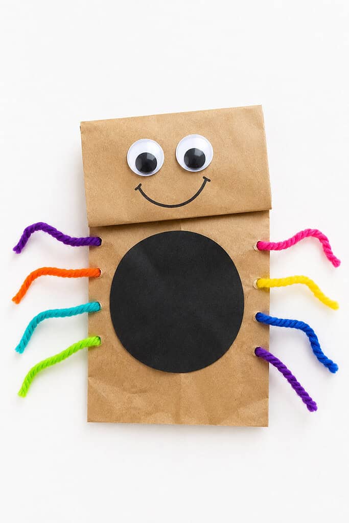 Brown paper bag spider puppet with googly eyes, a smiling face, a black paper circle body, and colorful yarn legs in purple, orange, teal, green, pink, yellow, and blue