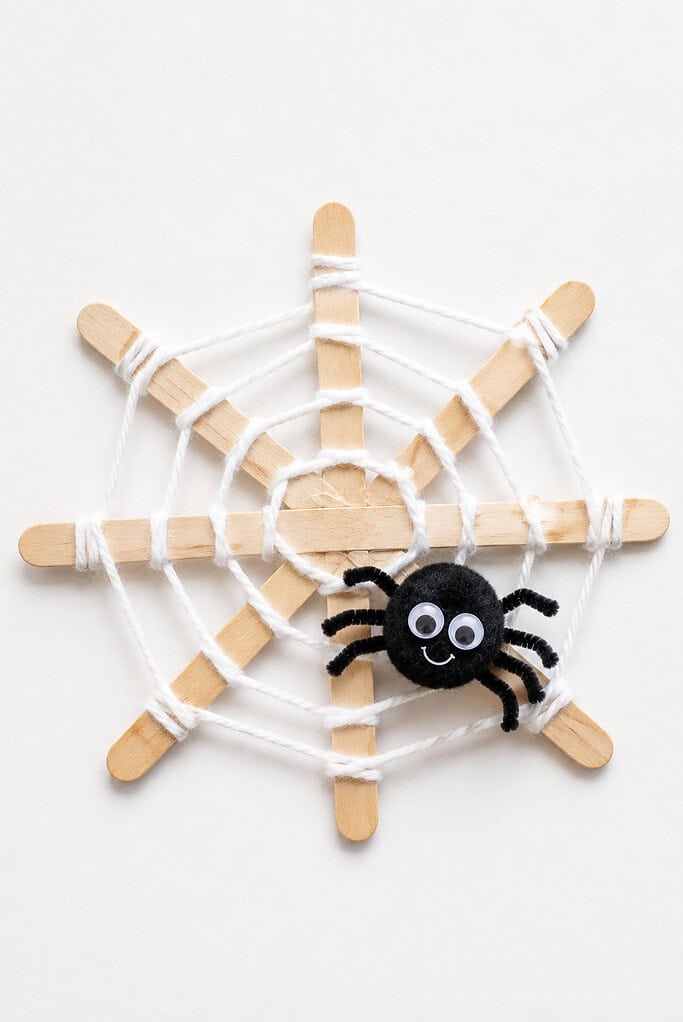 Wooden popsicle sticks arranged in a star shape and wrapped with white yarn to form a web, with a black pom-pom spider with googly eyes sitting in the middle