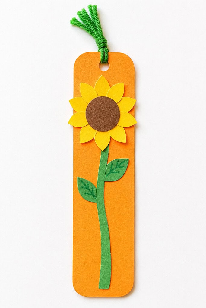 Orange cardstock bookmark with a yellow paper sunflower, brown center, green stem and leaves, and a green yarn tassel on a white table.
