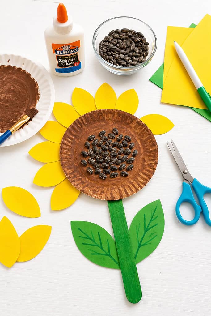 Flat lay of a kid's sunflower craft in progress on a white table with yellow paper petals, a painted paper plate, sunflower seeds, green paper leaves, glue, scissors, colored paper, a paintbrush, brown paint, and a marker.