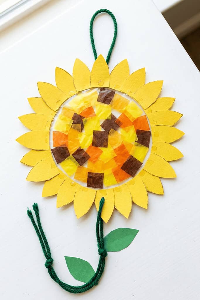 A child-friendly sunflower suncatcher craft made with yellow paper petals and colored tissue paper squares, resting on a white table.