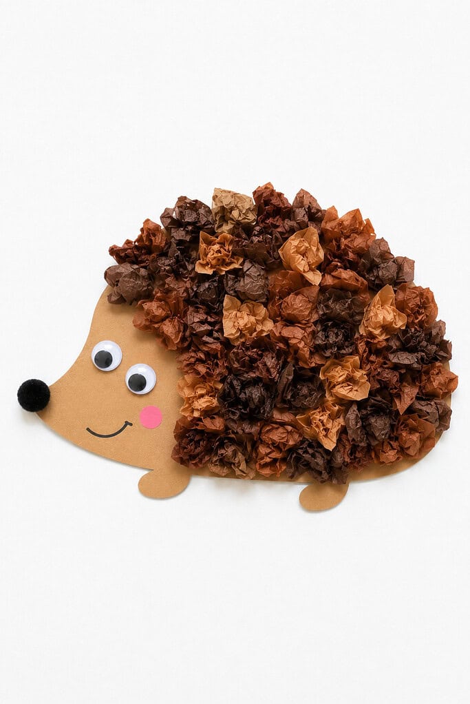 Small crumpled pieces of brown and tan tissue paper covering the back of a paper hedgehog for texture, with a tan face showing googly eyes, a black pom-pom nose, a pink cheek dot, and a smile