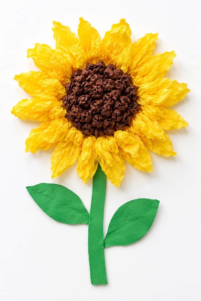 Yellow tissue paper sunflower craft with a crumpled brown center, green paper stem, and two leaves on a white table.