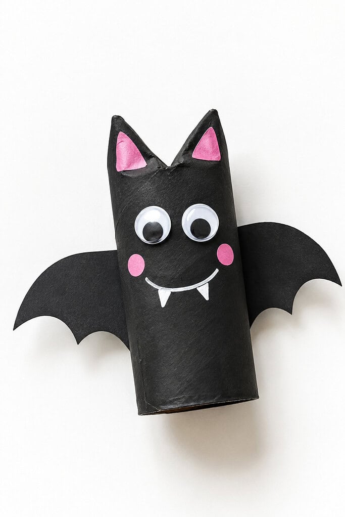 Black toilet paper roll bat craft with paper wings, pink ears and cheeks, googly eyes, a white smile, and small fangs on a bright white table.