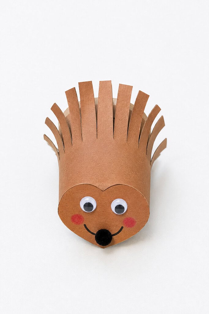 Brown cardboard tube with the top cut into fringe strips bent outward as spikes, over a heart-shaped tan paper face with googly eyes, a black nose, pink cheeks, and a smile