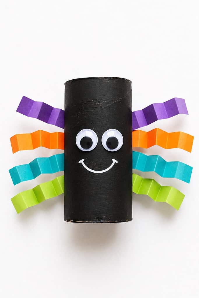 Black-painted toilet paper roll spider with googly eyes and a smile, and folded accordion-style paper legs in purple, orange, teal, and green