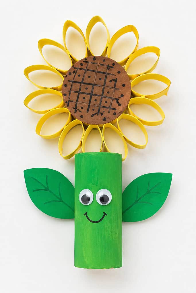 Green painted toilet paper roll with googly eyes, a smiling face, green paper leaves, yellow cardboard loop petals, and a brown paper center on a white table.