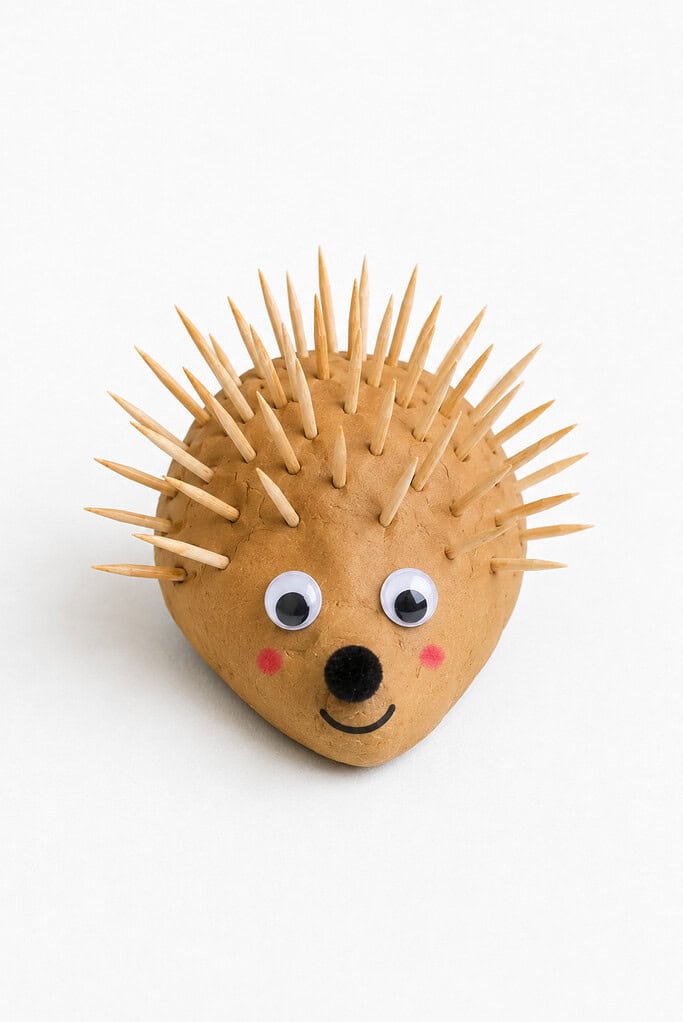 Ball of tan dough with wooden toothpicks pressed in as spikes, plus googly eyes, a black pom-pom nose, and pink cheeks on the front