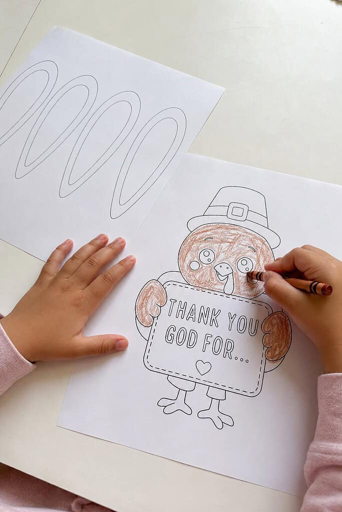 A child's hands coloring a black and white printed turkey page with a brown crayon on a white table, with a page of feather outlines beside it