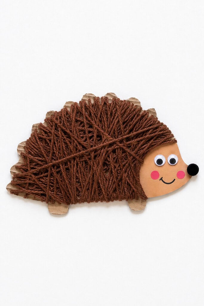 Brown yarn wrapped crisscross over a cardboard cutout body, attached to a tan paper face with googly eyes, pink cheeks, a black pom-pom nose, a smile, and small paper feet