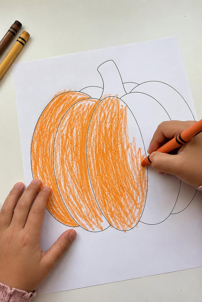 A child coloring the black-and-white printable pieces for a Thanksgiving pumpkin Sunday school craft with an orange crayon