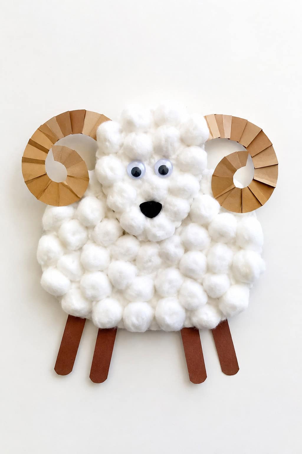 A fluffy cotton ball ram craft with curled paper horns, googly eyes, and paper legs on a white table.