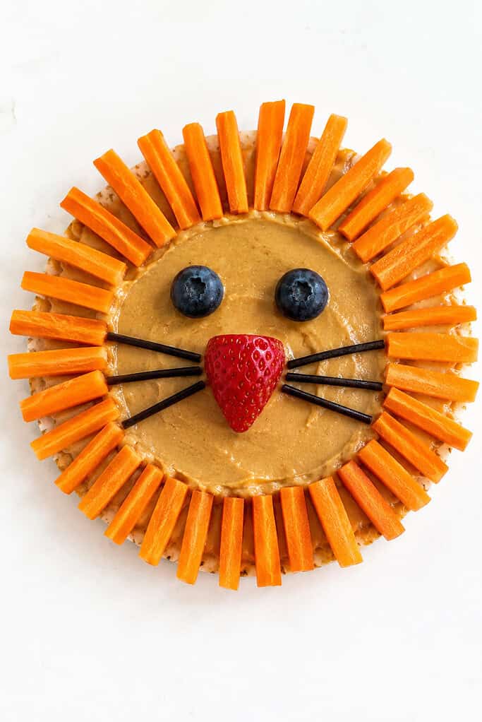 A graham cracker lion face with blueberry eyes, a strawberry nose, licorice whiskers, and a carrot mane.