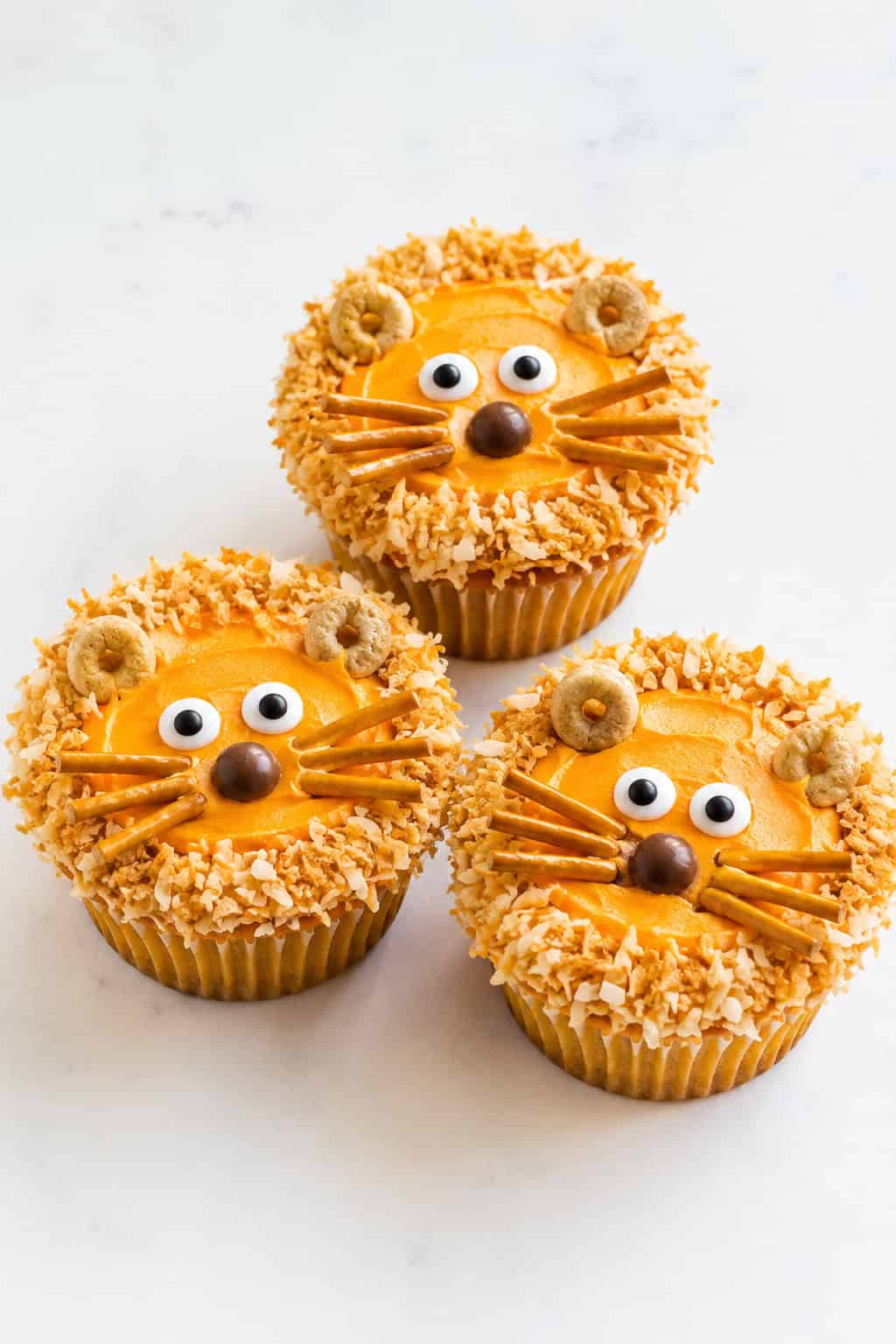 Three lion cupcakes with orange frosting faces, toasted-coconut manes, and pretzel whiskers on a white table.