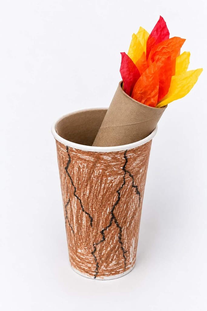 A white paper cup colored brown with crayon and drawn with black crack lines, with a toilet paper roll torch leaning inside it and red, orange, and yellow tissue-paper flames rising from the top.