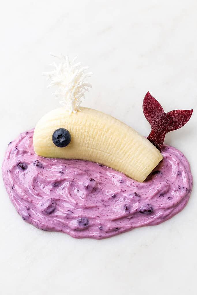 A banana shaped like a whale with a blueberry eye and fruit-leather tail on blue yogurt.