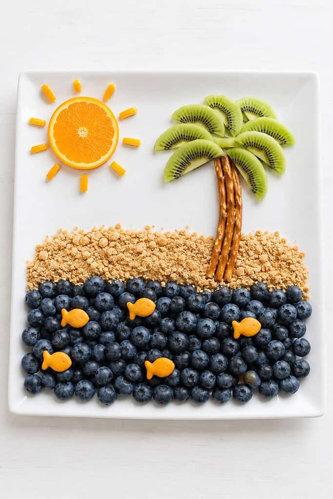 A plate with a kiwi palm tree, blueberry sea, graham-cracker sand, orange sun, and fish crackers.