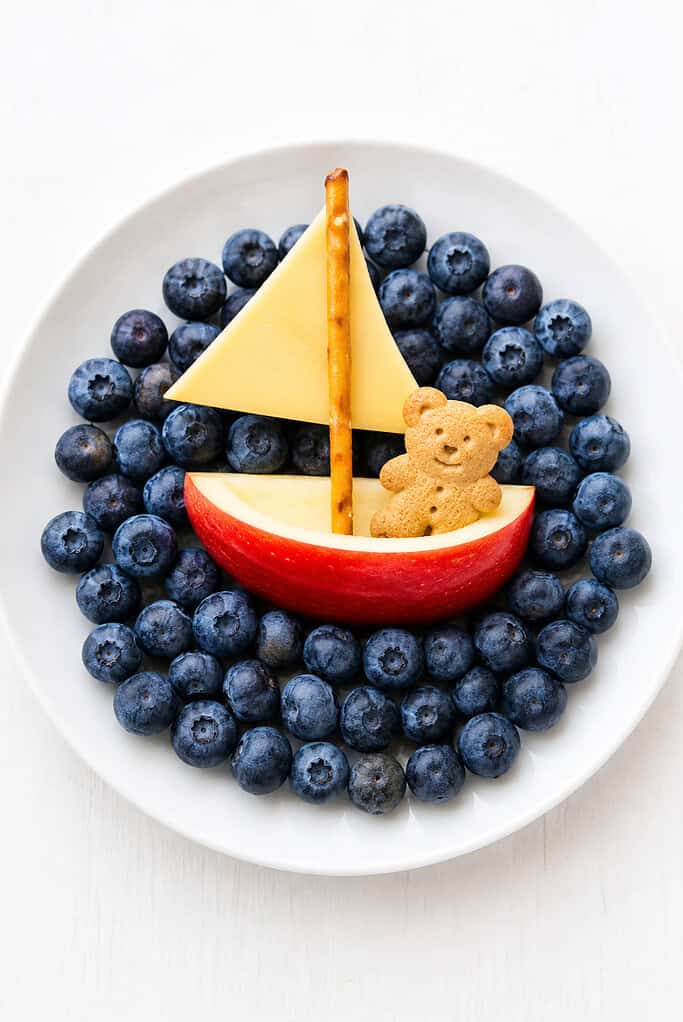 An apple-slice boat with a pretzel mast and cheese sail carrying a teddy-bear cracker on blueberries.