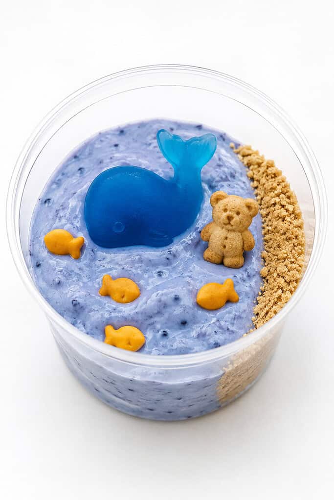 A clear cup with a blue yogurt sea, a whale gummy, a teddy-bear cracker, and fish crackers.