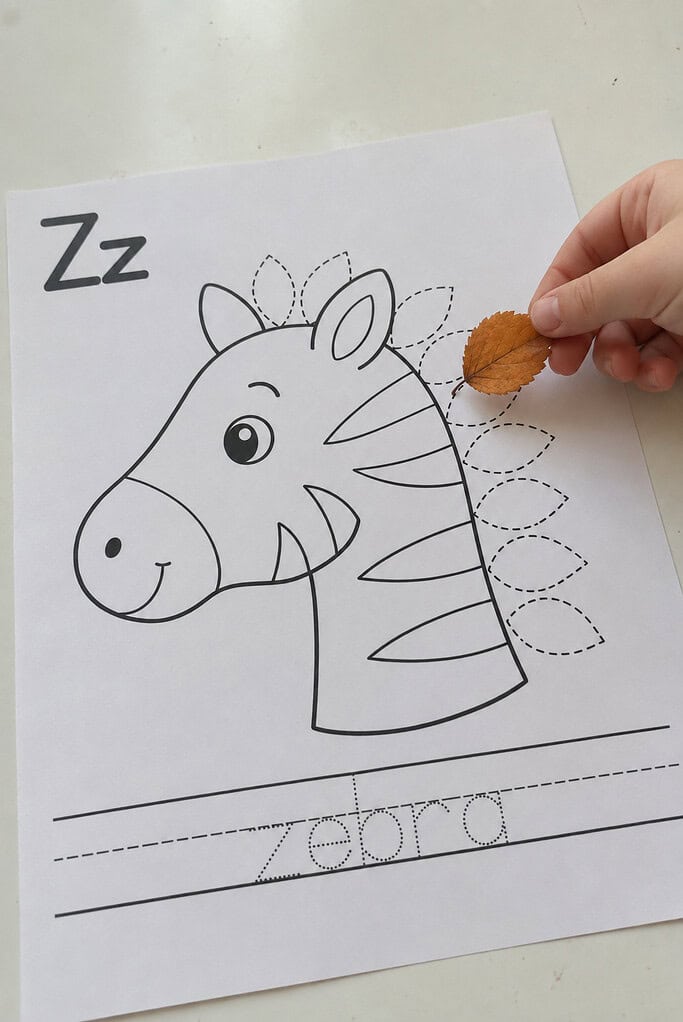 A child holding a real leaf above the dashed guide on a leaf alphabet craft worksheet to match the shape