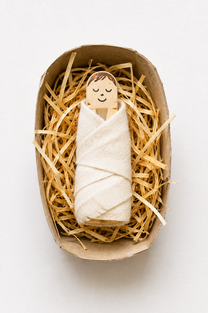 A swaddled clothespin baby Jesus with a sleeping face resting in a half-tube manger filled with straw.