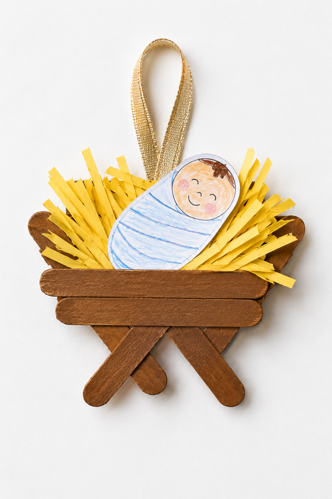 A popsicle stick manger with yellow paper hay, a swaddled baby Jesus, and a ribbon loop for hanging.