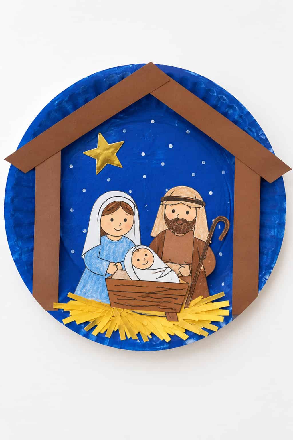 A paper plate nativity with a painted night sky, a stable roof, and cut-out Mary, Joseph, and baby Jesus.