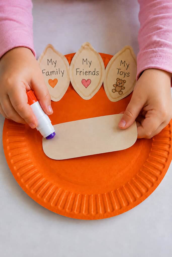 A child gluing the "Thank You God For" sign onto the paper plate pumpkin Thanksgiving Bible craft