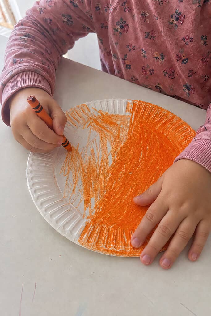 A child coloring a paper plate orange to make the pumpkin for a paper plate pumpkin Thanksgiving Bible craft
