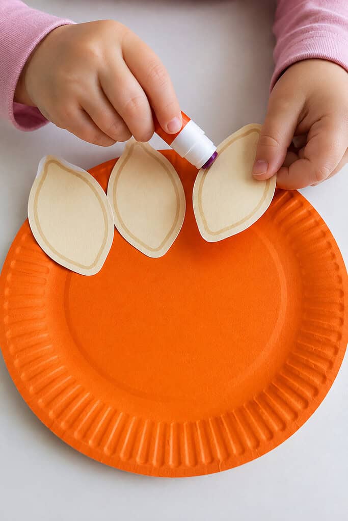 A child gluing thankful pieces across the top of the orange paper plate for a paper plate pumpkin Thanksgiving Bible craft