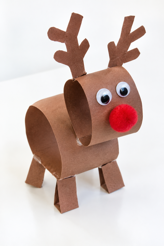 a 3D paper reindeer made of brown paper loops standing on folded legs with antlers, googly eyes, and a red pom-pom nose on a white table