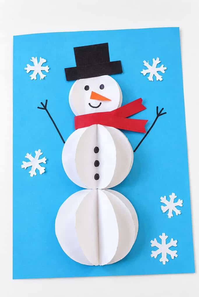 a 3D paper snowman craft made of folded white paper balls on blue paper with a black hat, red scarf, dot eyes, and stick arms on a white table