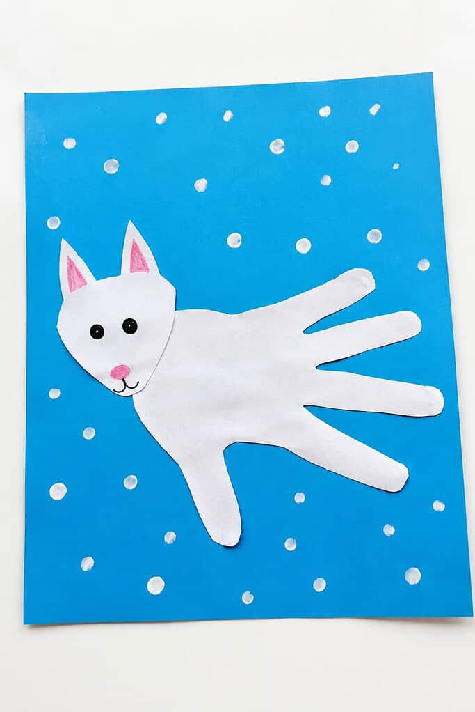 a white handprint arctic fox craft with a pointed-ear head, pink nose, and bushy fingered tail on a light blue snowy background on a white table