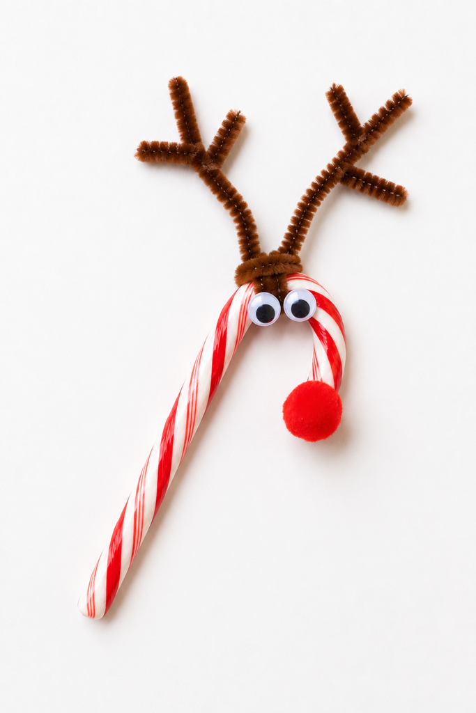 a candy cane reindeer craft made from a red and white candy cane with brown pipe cleaner antlers, googly eyes, and a red pom-pom nose on a white table