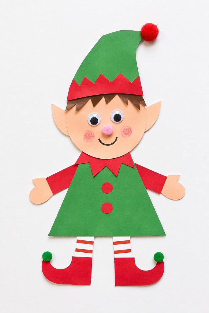 a paper Christmas elf craft with a round head, a pointed pom-pom hat, googly eyes, rosy cheeks, a triangle body, little arms, and pointy shoes on a white table