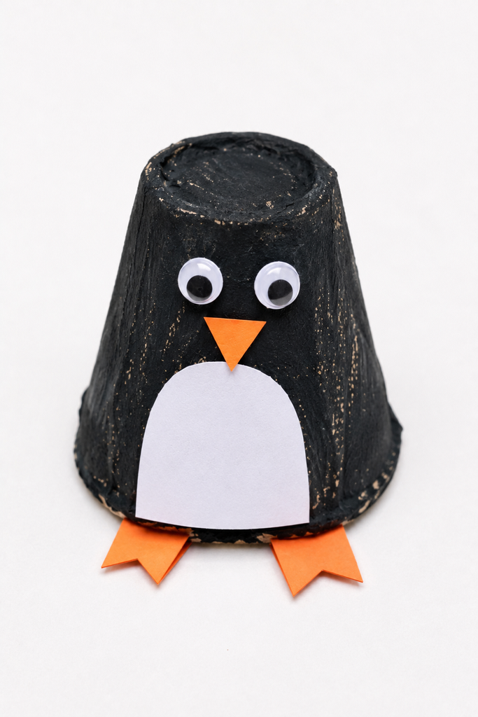 a standing egg carton penguin painted black with a white belly, googly eyes, orange beak, and orange feet