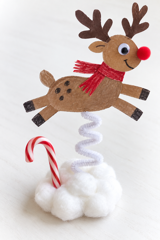 a paper reindeer on a coiled pipe cleaner spring bobbing over a mound of cotton snow with a candy cane on a white table