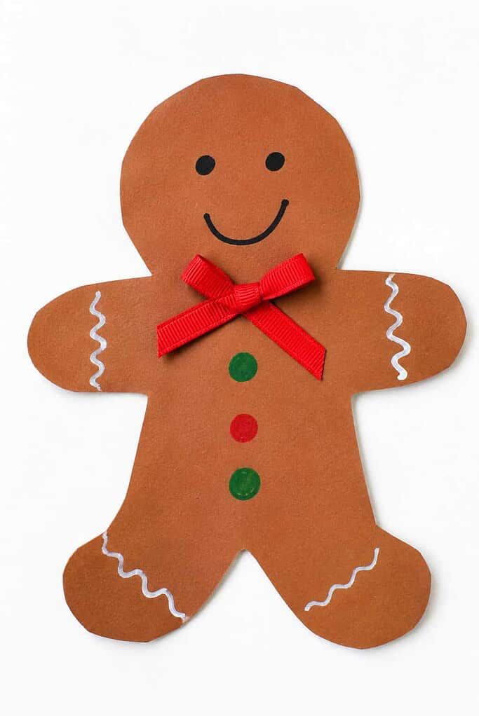 a brown paper gingerbread man craft with dot eyes, a smile, button dots, white icing lines on the arms and legs, and a ribbon bow on a white table