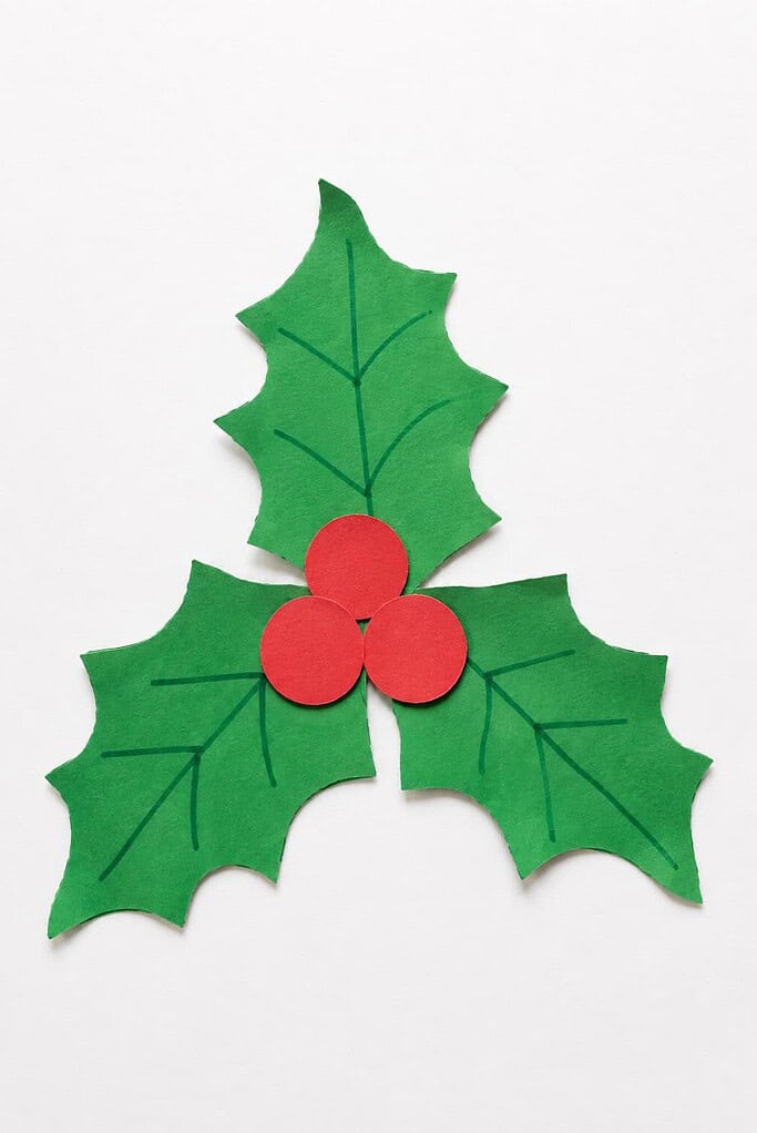 a holly craft made of three green paper leaves with pointed edges and three red paper berries in the center on a white table