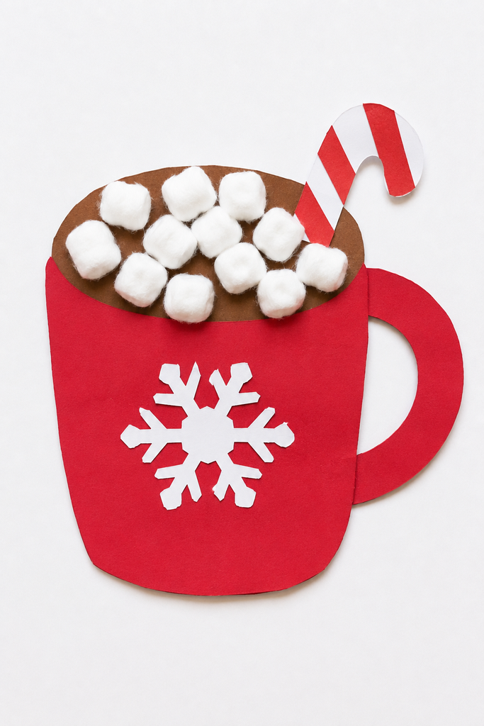 a paper hot chocolate mug craft with a handle, cotton ball marshmallows spilling over the rim, a snowflake, and a paper candy cane on a white table