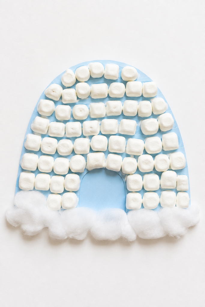 a marshmallow igloo craft made from a paper dome covered in rows of mini marshmallows with an open arched doorway, sitting on cotton snow on a white table