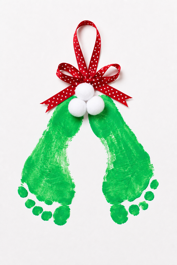 a mistletoe footprint craft made from two green painted footprints as leaves with a cluster of berries, a ribbon bow, and a hanging loop at the top on a white table