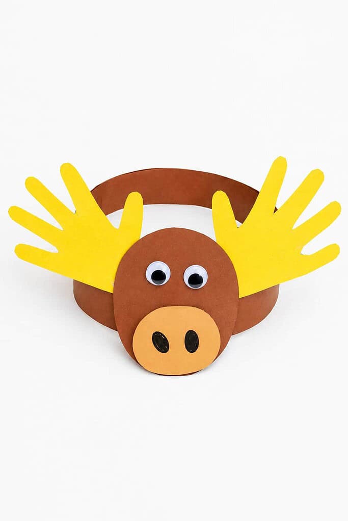 a moose headband craft made from a brown paper band with a moose face, googly eyes, an oval snout, and two handprint antlers on a white table