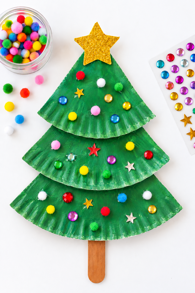 A folded green paper plate Christmas tree with gems and pom-poms beside a dish of pom-poms.