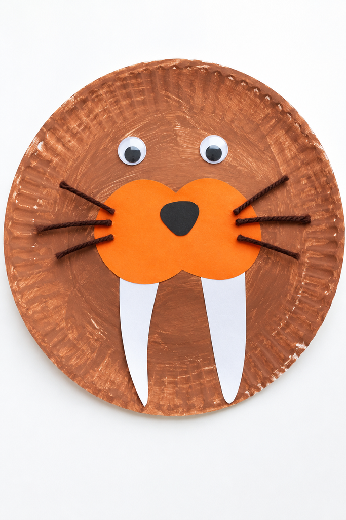 a brown paper plate walrus craft with googly eyes, an orange snout, a black nose, yarn whiskers, and two white paper tusks on a white table
